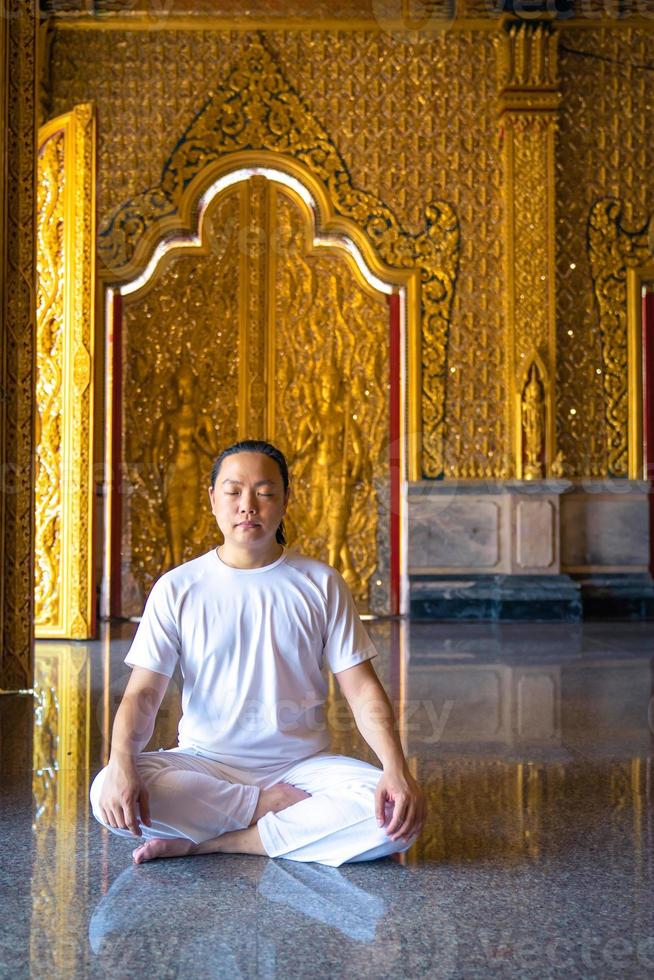 aziatische langharige man ontspant meditatie met een geheel wit kostuum zit voor het gouden behang van de boeddhist in de tempel, thailand. foto