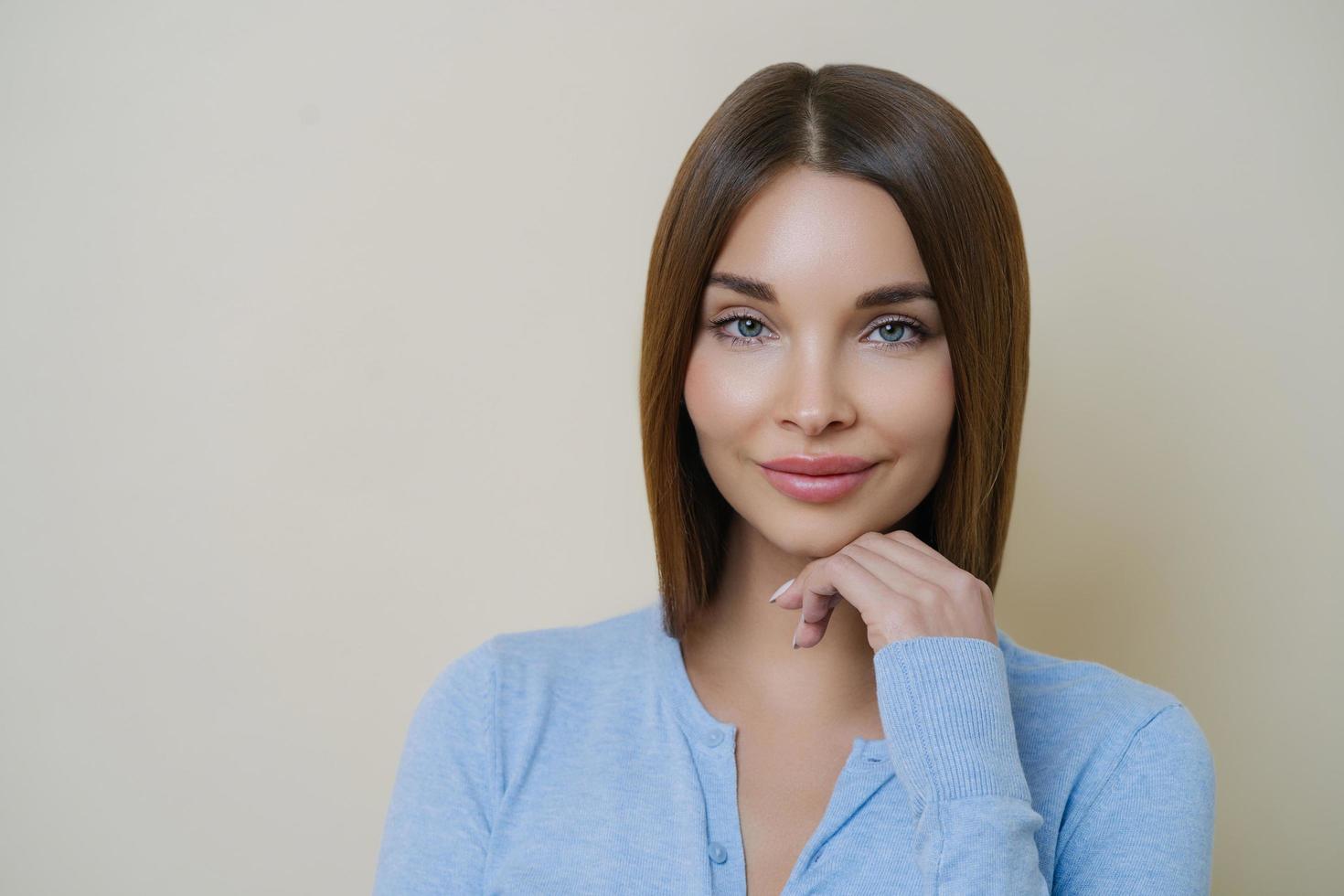 portret van mooie donkerharige vrouw met een gezonde natuurlijke schone huid, raakt kin aan, heeft manicure en draagt blauwe trui, geïsoleerd op beige achtergrond. mensen, schoonheid, gezichtsuitdrukkingen concept. foto