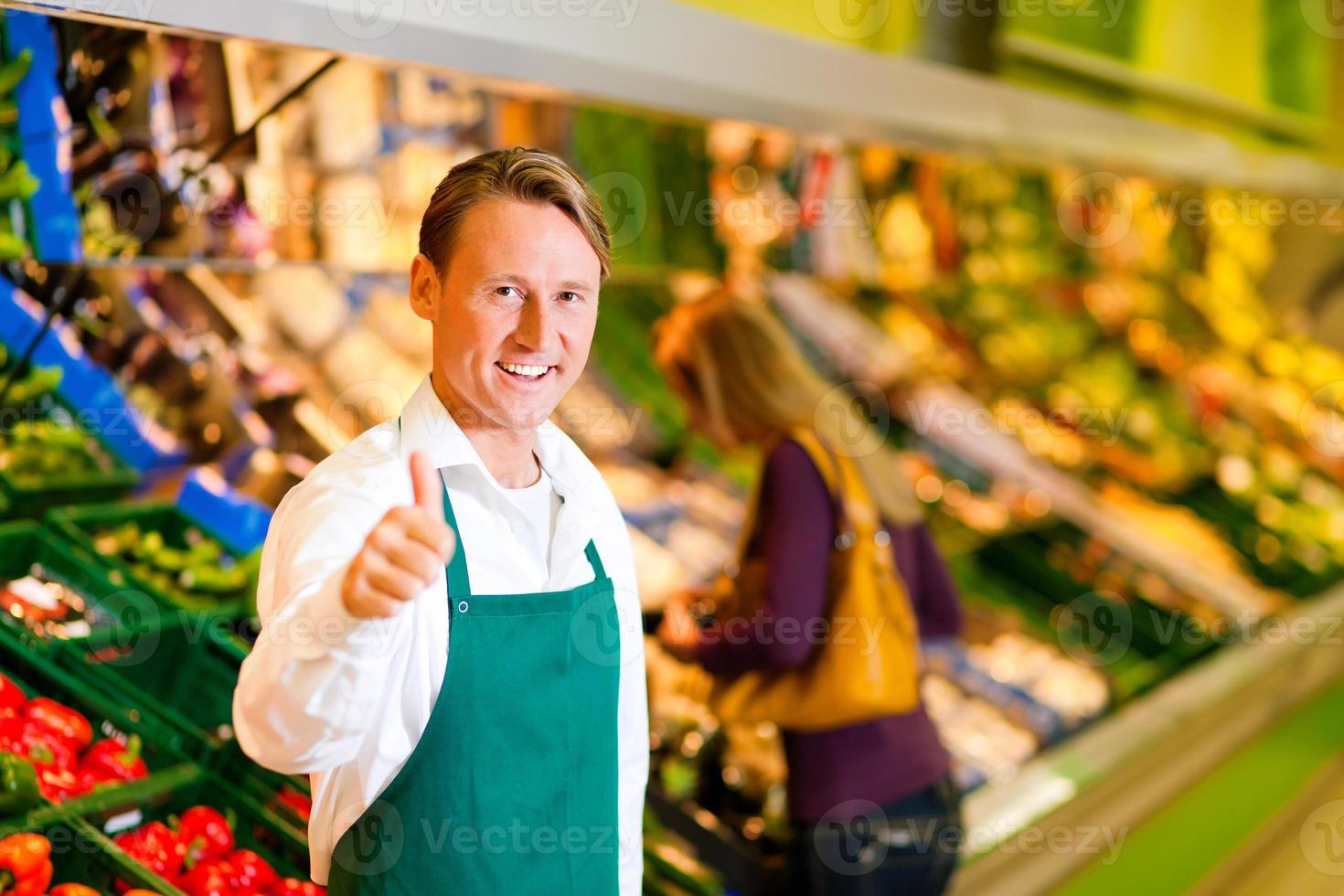 glimlachende mens in groene schort met één duim omhoog bij supermarkt foto