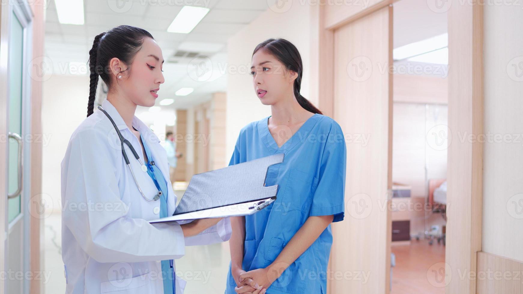 professionele, Aziatische jonge vrouwelijke arts, personeel in uniform staande in ziekenhuisgang. twee collega-werknemers bespreken, werken samen met medisch rapportklembord op de gang van de kliniek. foto
