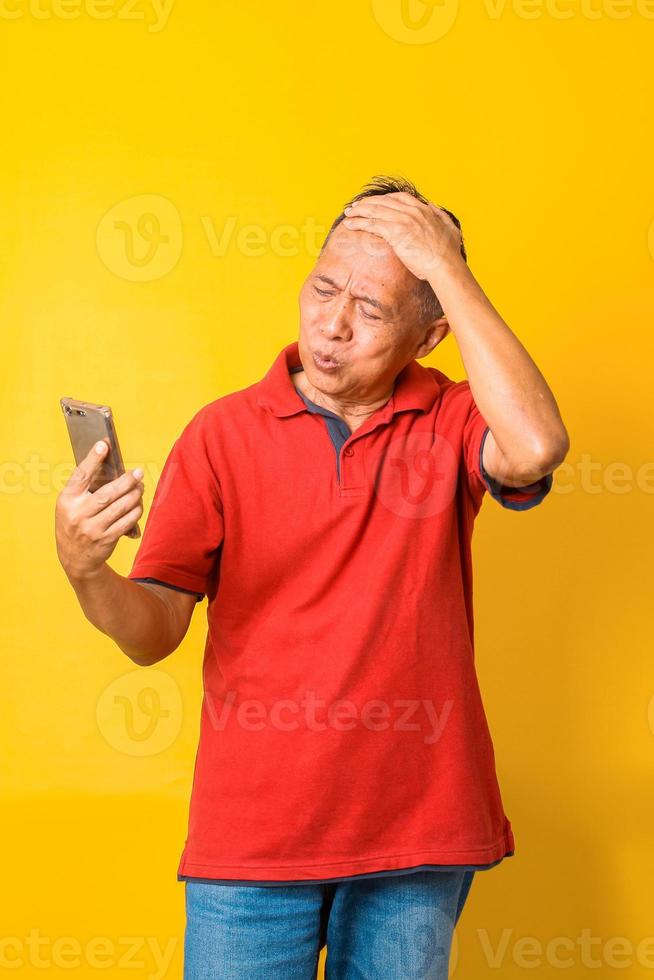 Aziatische senior man op zoek naar smartphone gestrest met de hand op het hoofd, geschokt met verrassingsgezicht, boos en gefrustreerd, geïsoleerd op gele achtergrond. angst en boos voor fouten. foto