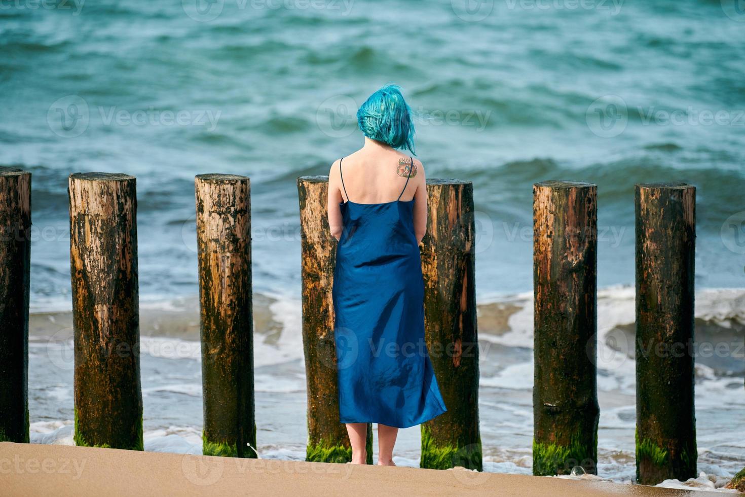 jonge blauwharige vrouw in lange donkerblauwe jurk die op het zandstrand staat en naar de zeehorizon kijkt foto