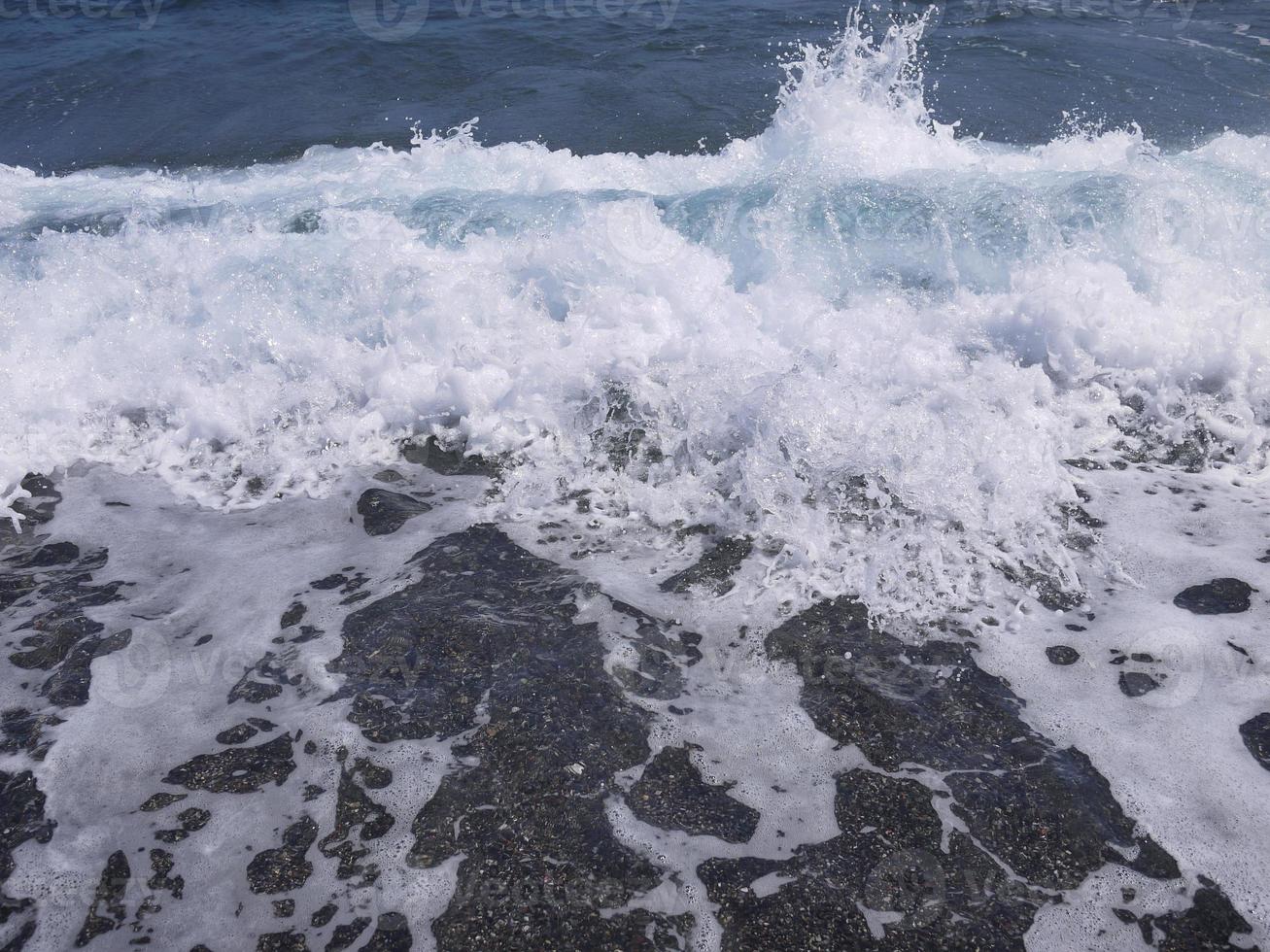 diepblauwe zee op zwart strand met witte plonsgolf voor achtergrondbehang foto