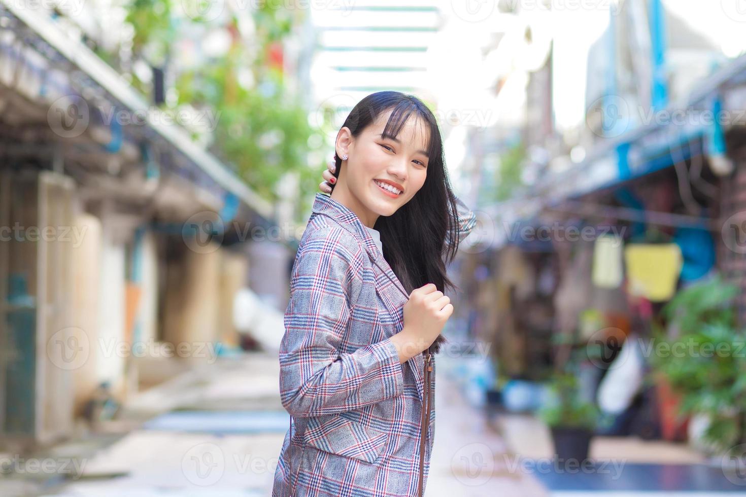 zelfverzekerde jonge Aziatische vrouw die een grijs gestreepte blazer en schoudertas draagt, lacht vrolijk en kijkt naar de camera terwijl ze door de oude stad naar haar werk pendelt. foto