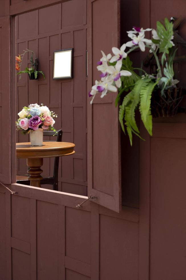 zijaanzicht en selectieve focus op kunstmatige roze bloemen in keramische vaas op houten ronde tafel met lege fotolijst op houten muur in raamkozijn en wazige voorgrond, huisdecoratie concept foto