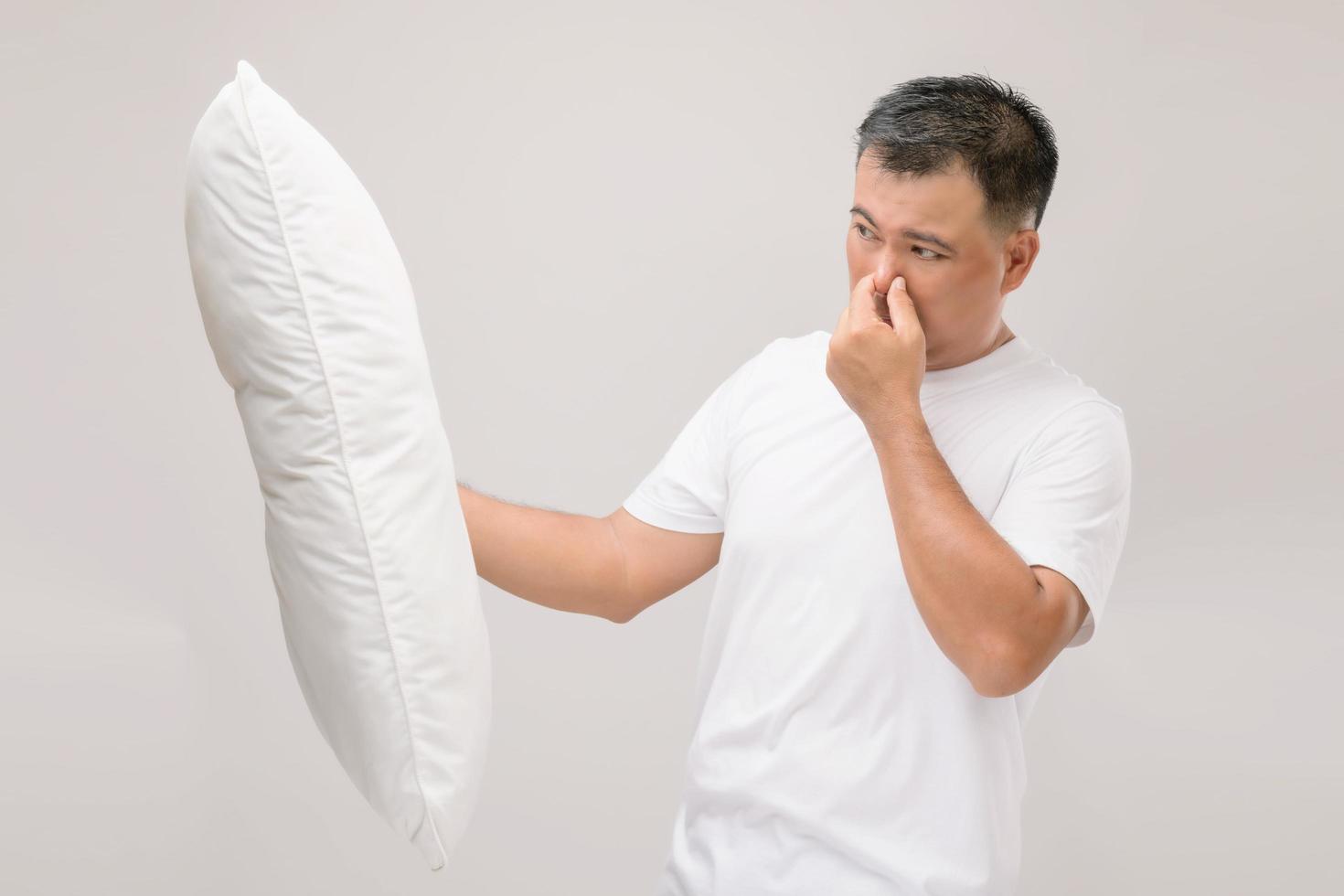 het kussen stinkt. portret aziatische man die wit kussen vasthoudt en een slechte geur krijgt. studio-opname op grijs foto