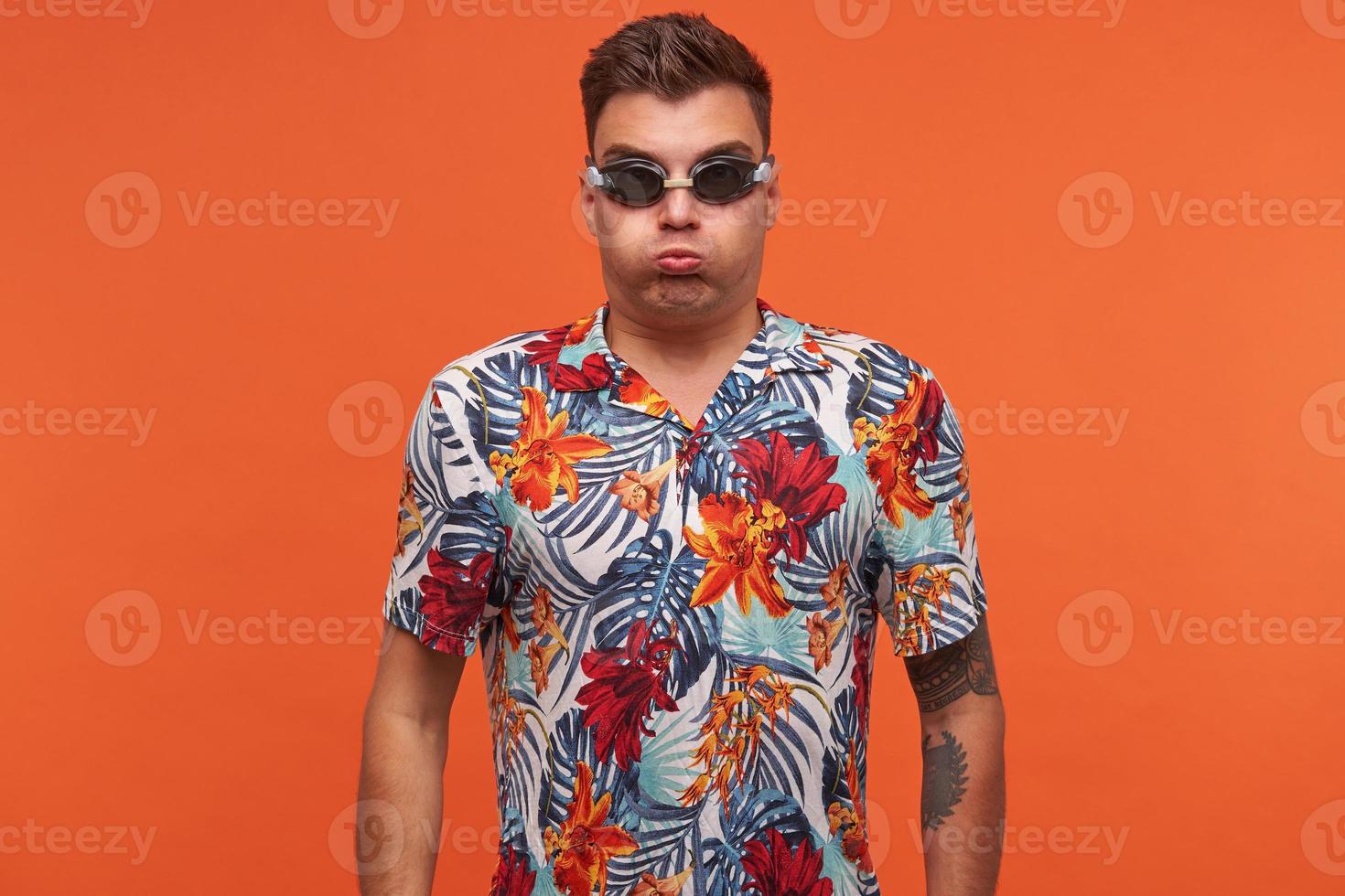 studio-opname van een jonge, aantrekkelijke man die over een oranje achtergrond staat met zijn handen naar beneden, lucht in zijn mond neemt en adem inhoudt, een zwembril draagt foto