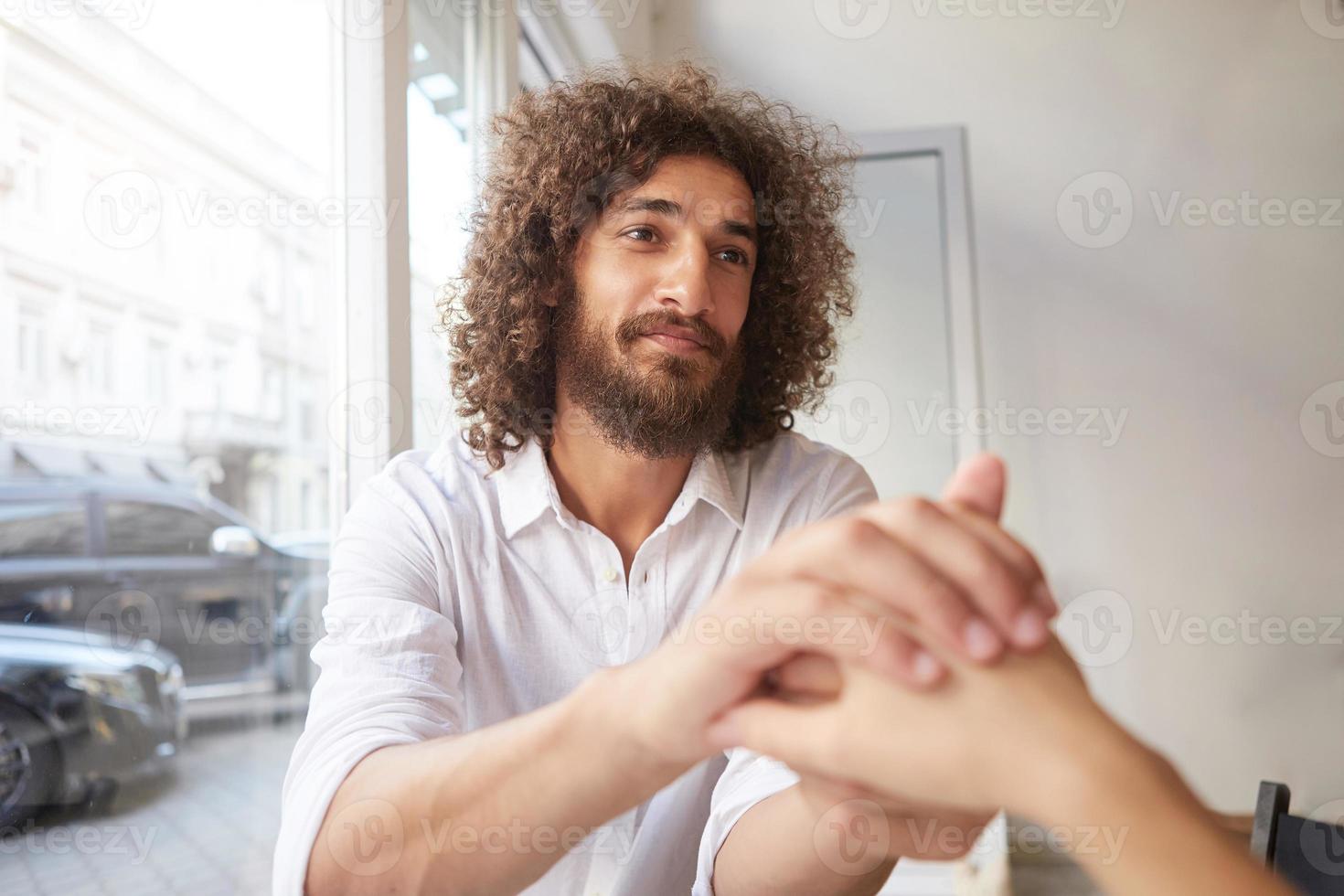 Jonge aantrekkelijke gekrulde man met baard en bruine vriendelijke ogen die bij het raam zit, vrouwelijke hand vasthoudt en haar innig aankijkt, wit overhemd draagt foto