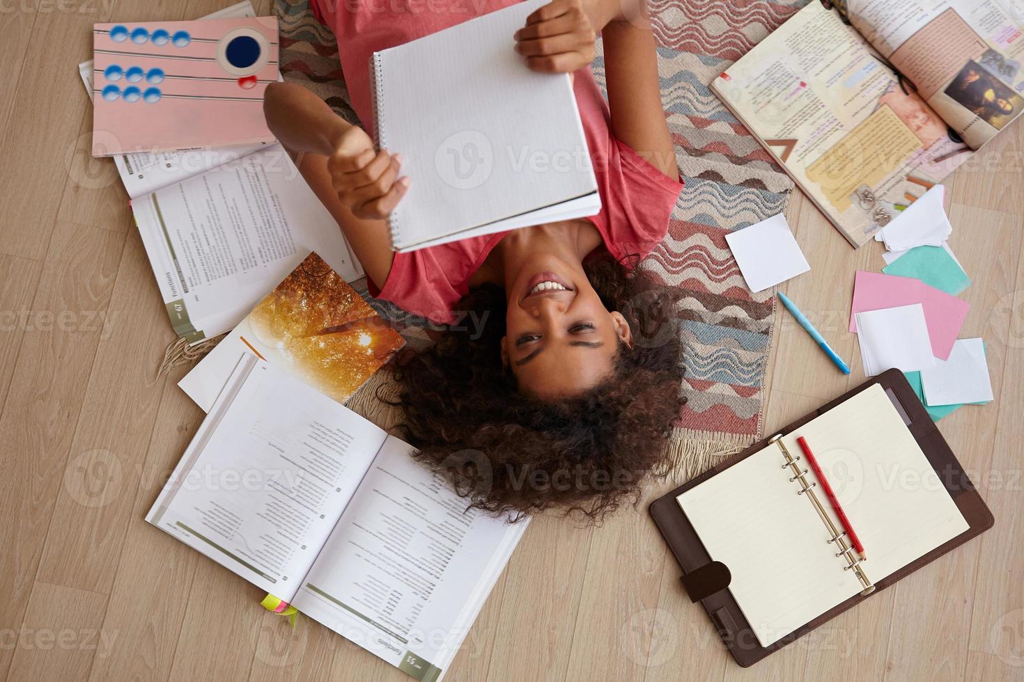 bovenaanzicht van aantrekkelijke jonge krullende vrouw met donkere huid liggend op tapijt tijdens het lezen van notities, voorbereiding op examens met veel boeken, roze t-shirt dragen foto