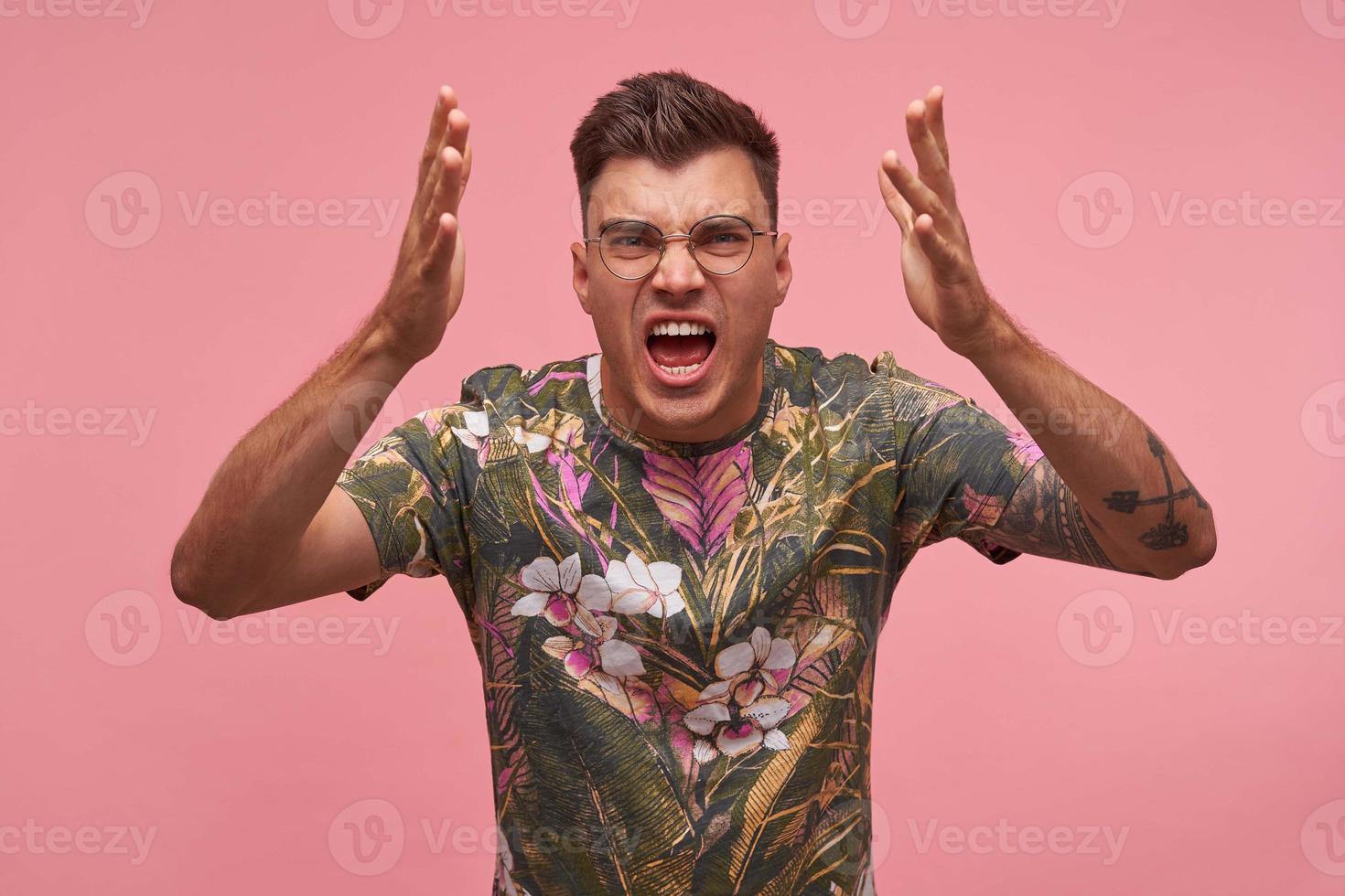 gekke knappe jonge donkerharige man die over een roze achtergrond staat met wijd opengesperde mond, zijn handen gewelddadig opheffend en schreeuwend van woede foto