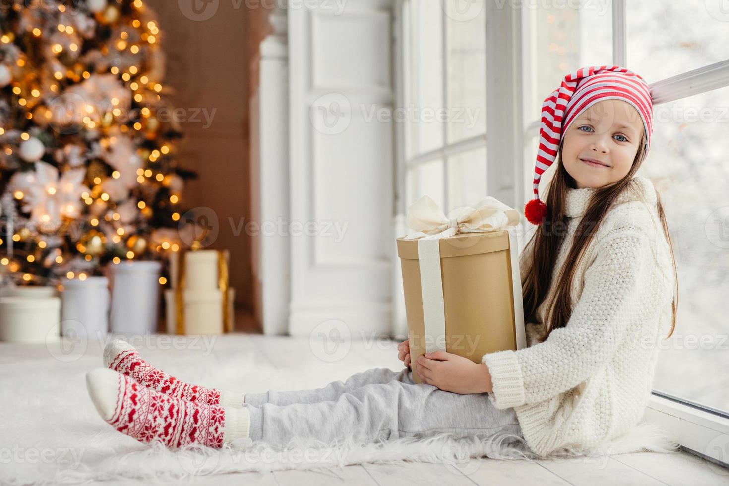 schattig mooi meisje met cadeau, zit in de buurt van raam in woonkamer, kijkt heerlijk in de camera, bewondert prachtig mooi versierde dennenboom, heeft een goed humeur en vakantie. nieuwjaarsconcept foto