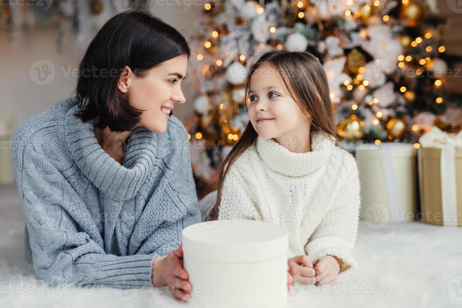 mensen, familie, feest en vakantie concept. moeder en dochter met aantrekkelijk uiterlijk kijken elkaar aan, liggen op warm wit tapijt in de buurt van versierde nieuwjaarsboom en geschenkdoos foto