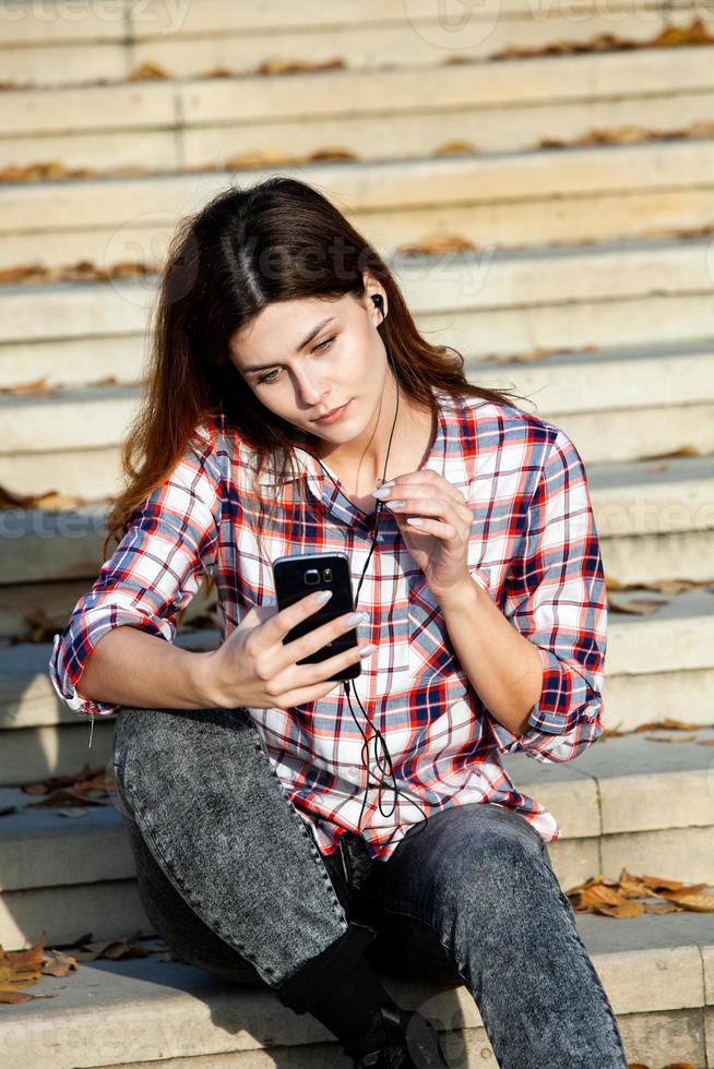 afbeelding van mooie stijlvolle vrouw met mobiele telefoon. jong europees meisje dat op straat staat en een mobiele telefoon gebruikt. vrouw die naar muziek luistert met de telefoon en plezier heeft. mobiel internetconcept. foto