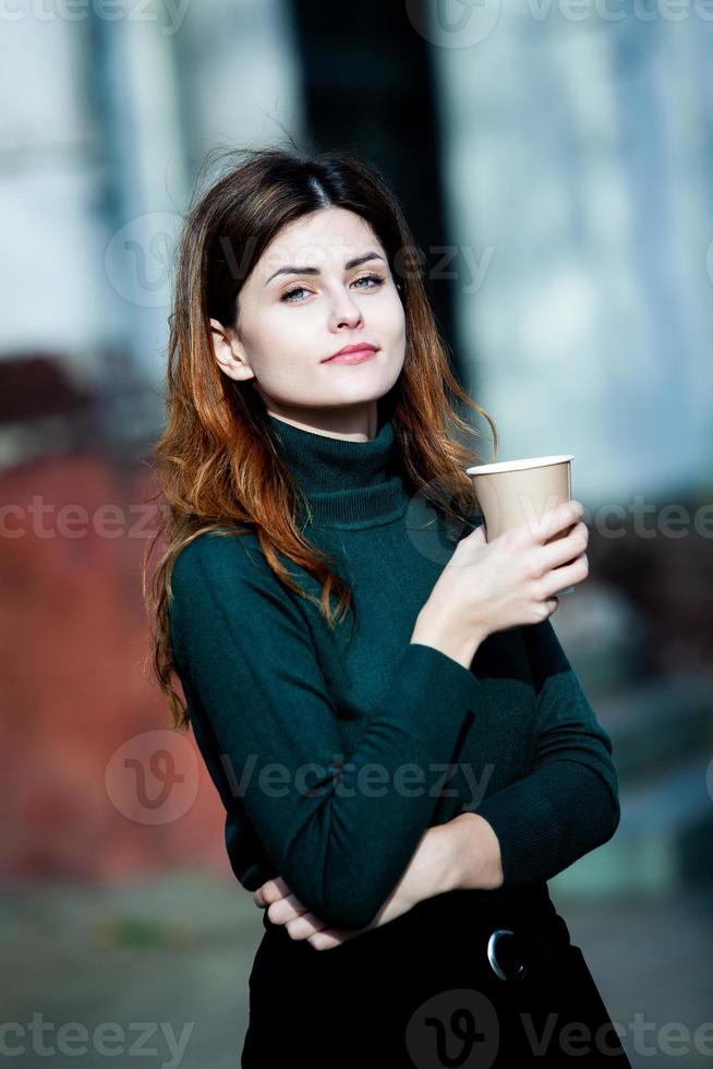 jonge stijlvolle vrouw die thee drinkt in een stadsstraat. hipster europees meisje met latte papier glas. prachtige jonge vrouw met kopje koffie in de stad straat. koffiepauze. koffie om mee te nemen. foto