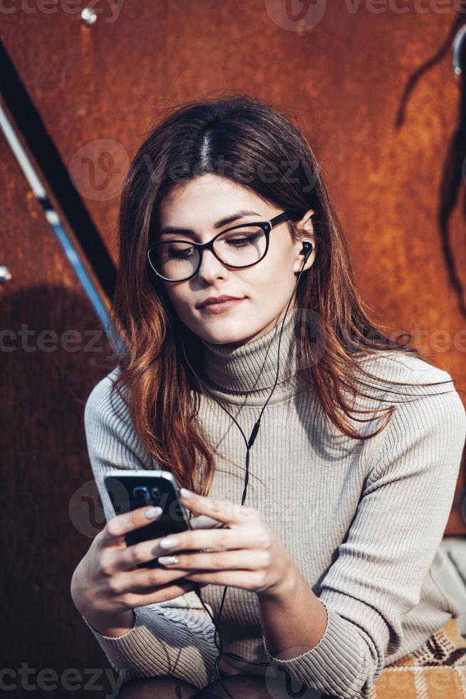 afbeelding van mooie stijlvolle vrouw met mobiele telefoon. jong europees meisje dat op straat staat en een mobiele telefoon gebruikt. vrouw die naar muziek luistert met de telefoon en plezier heeft. mobiel internetconcept. foto