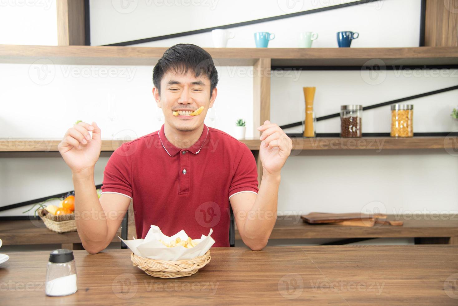 aziatische jonge man met een casual rood t-shirt geniet van het ontbijt, het eten van frietjes met een lach. jonge man die eten kookt in de keukenkamer in loftstijl foto