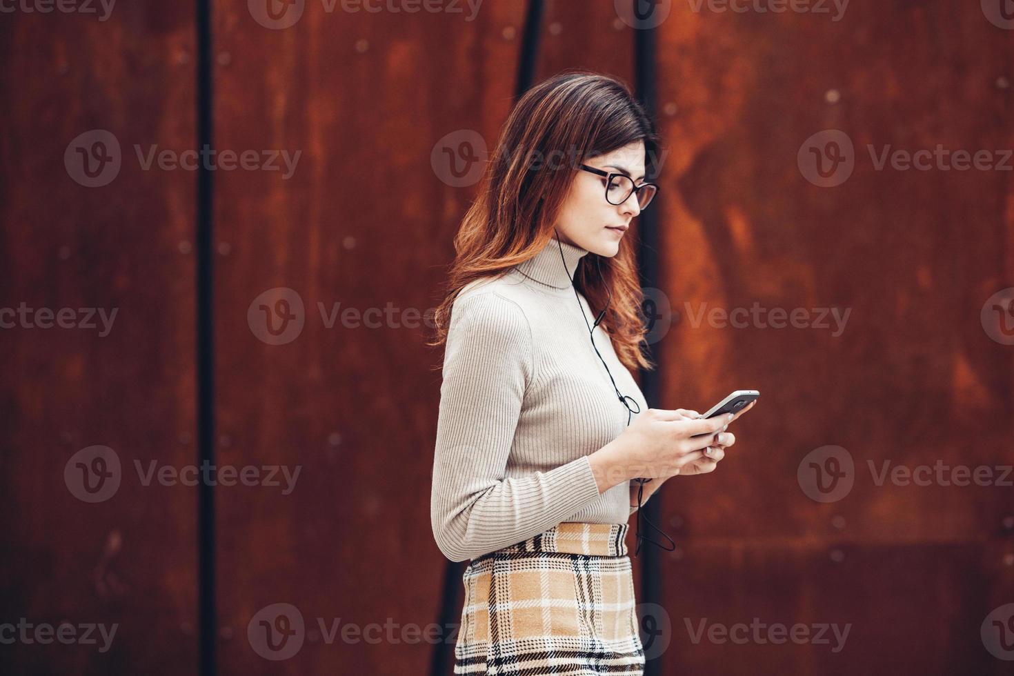 afbeelding van mooie stijlvolle vrouw met mobiele telefoon. jong europees meisje dat op straat staat en een mobiele telefoon gebruikt. vrouw die naar muziek luistert met de telefoon en plezier heeft. mobiel internetconcept. foto