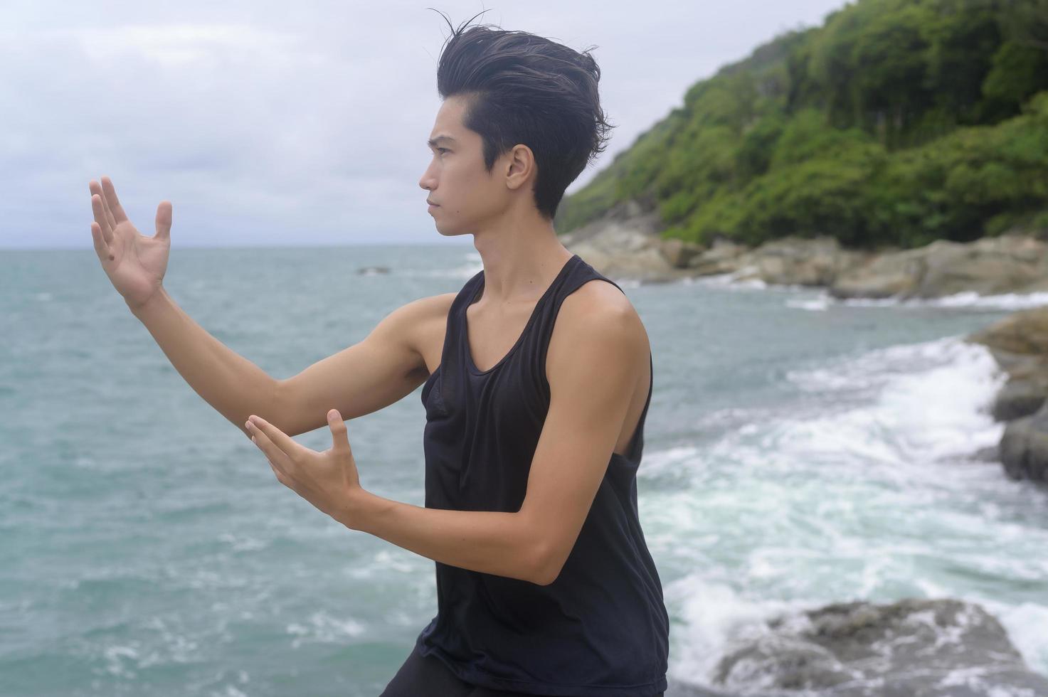 jonge man in sportkleding die vechtsport, qigong, tai chi op de rots aan zee doet, gezondheids- en meditatieconcept foto