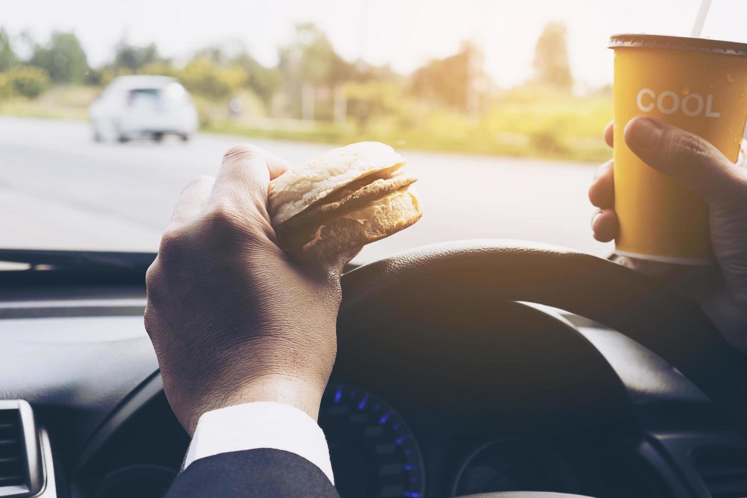 man autorijden terwijl hij een kopje koude koffie vasthoudt en hamburger eet foto