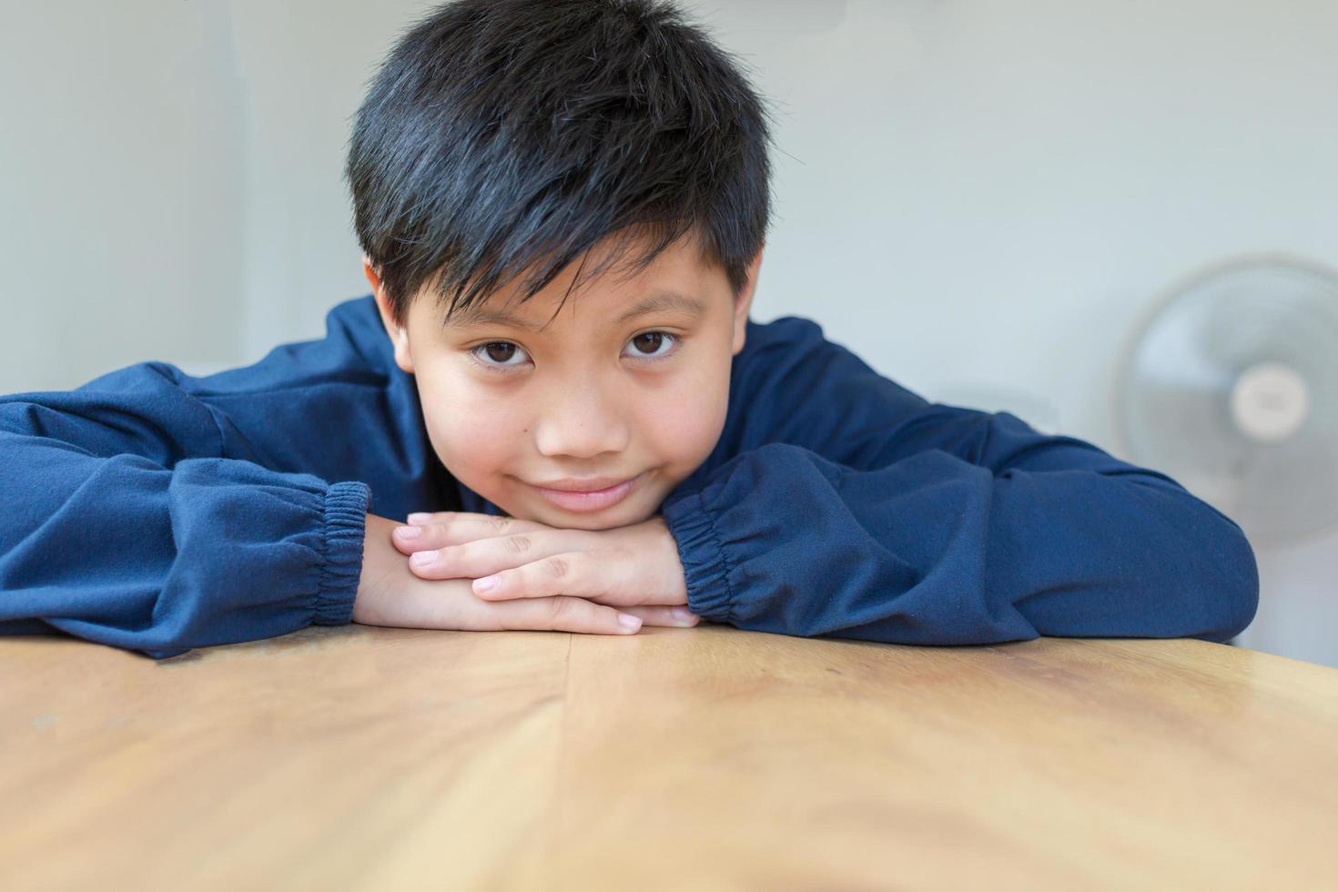 schattige aziatische jongen met een blanke huid die lacht en in de camera kijkt en gelukkig kinnen op hands-on houten tafel laat rusten. close-up portret van een schattig kind foto