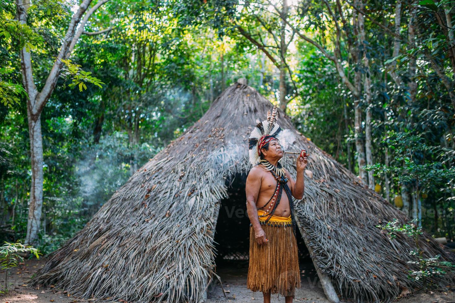 sjamaan van de pataxo-stam, gekleed in veren hoofdtooi en pijp rokend. braziliaanse indiaan die naar rechts kijkt foto