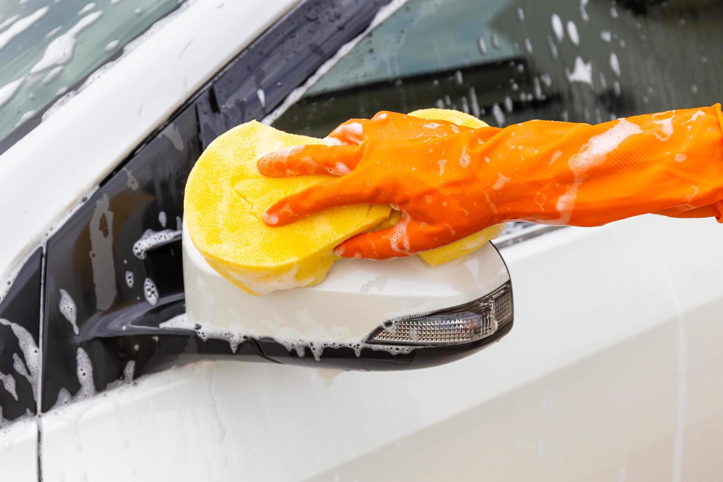 vrouwenhand die oranje handschoenen met gele spons draagt die zijspiegel moderne auto wassen of auto schoonmaken. carwash concept foto