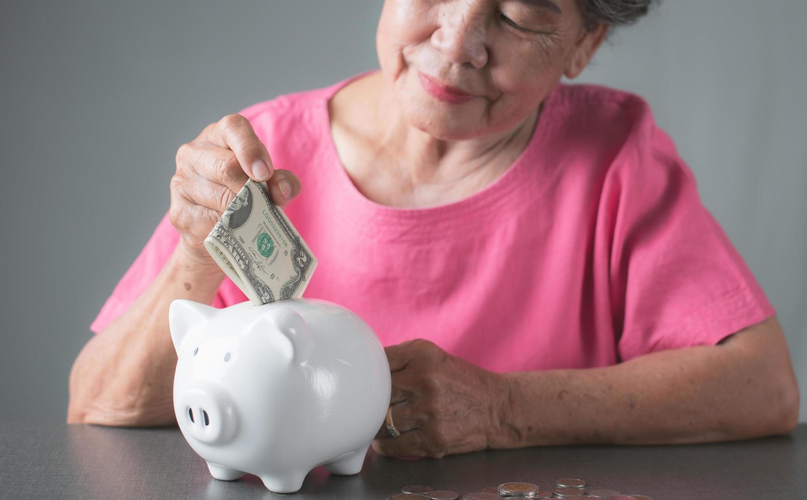 oude senior vrouw dollarbiljetten in een wit spaarvarken op tafel zetten. foto
