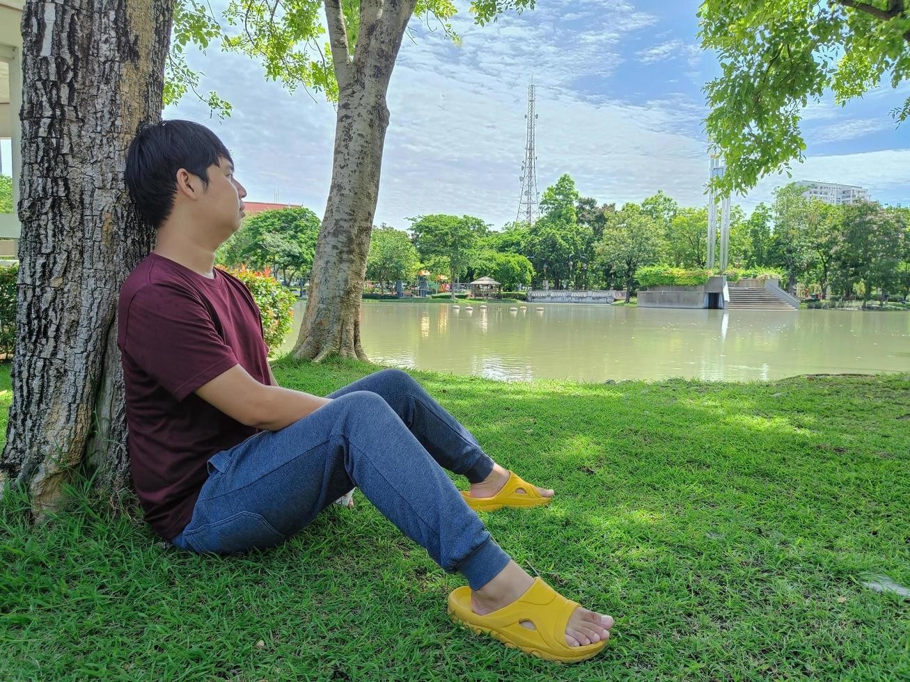 een jonge aziatische man met zwart haar zit onder een boom en kijkt naar de lucht en de wolken. stress en angst. wat denk je in de zomerparkstad? foto