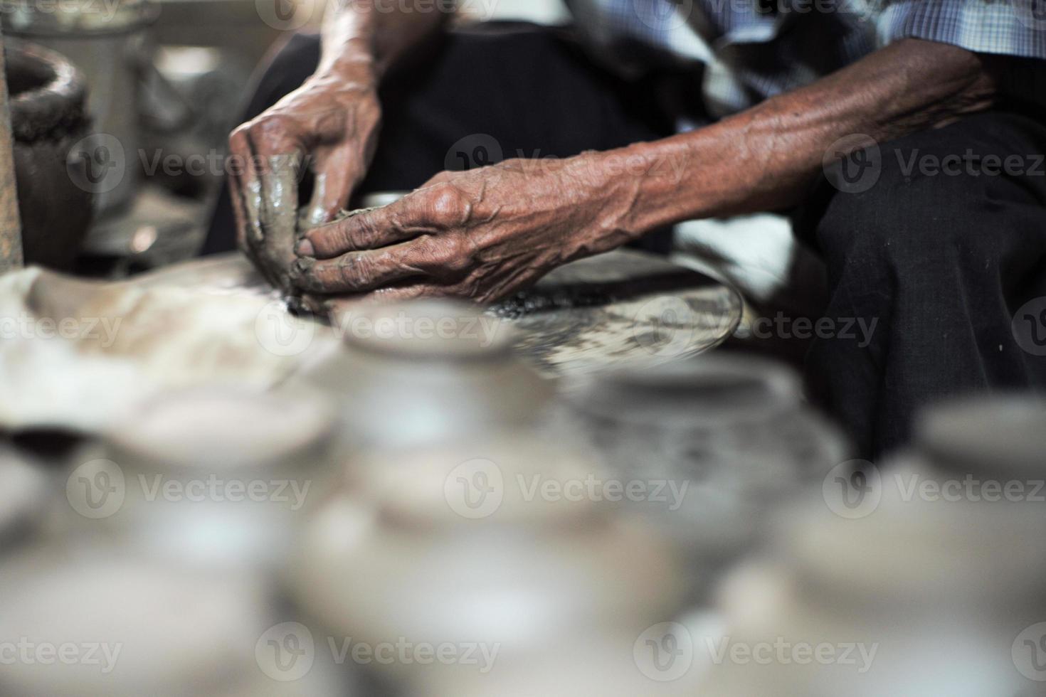 selectief gefocust op de vuile gerimpelde huid handen van oude man die de klei aan het vormen is werken op het spinnewiel voor het maken van de pot met wazige groep kleipotten op de voorgrond foto