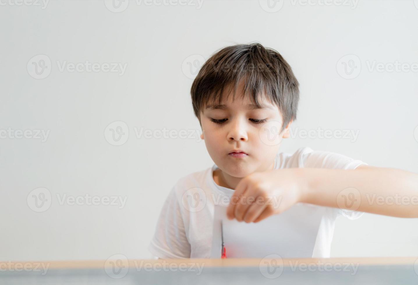 kind met een schaar die wit papier snijdt, schooljongen die kunst en ambacht maakt voor zijn huiswerk, kind leert hoe de schaar te gebruiken, snijd het papier, kinderen leren en spelen thuis, thuisschoolconcept foto