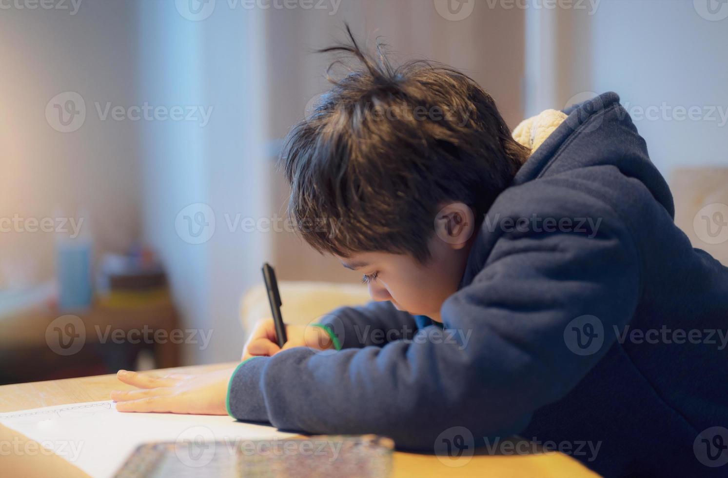 kind dat op tafel zit en huiswerk doet, kindjongen met zwarte pen die op wit papier schrijft, jonge jongen die Engelse woorden thuis oefent. basisschool en thuisonderwijs, concept voor afstandsonderwijs foto