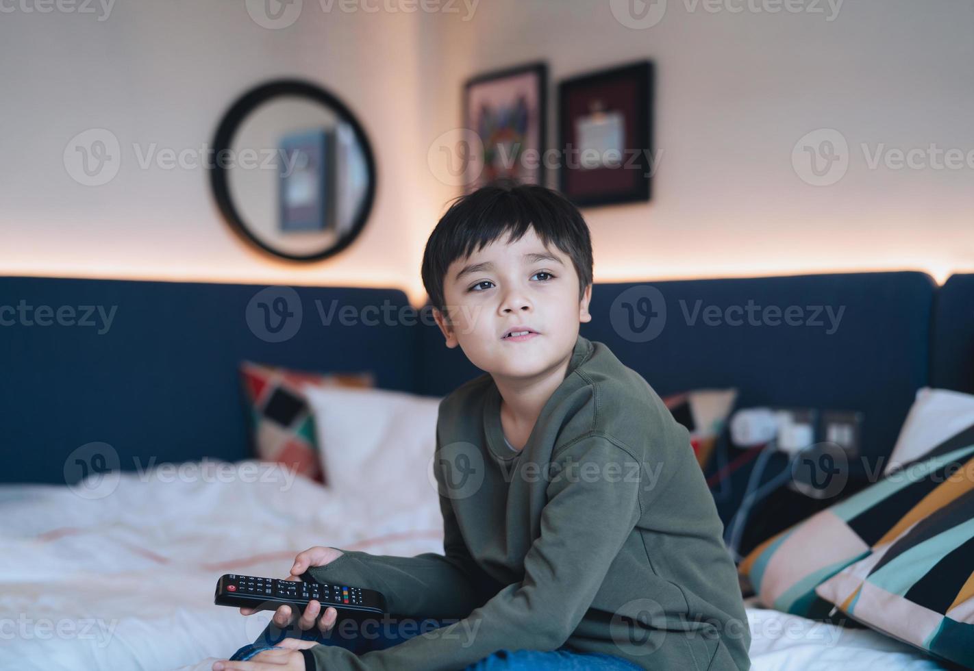 gelukkige jonge jongen zittend in bed met afstandsbediening, schattige jongen kijkt uit met lachend gezicht, kind ontspannen thuis tv kijken in bed kamer. foto