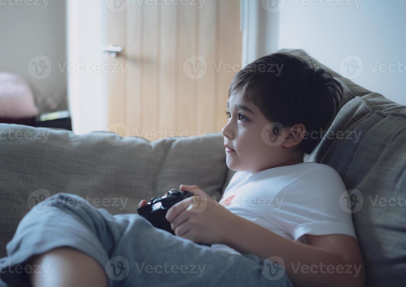 gelukkige jonge jongen die online videogames speelt met vrienden, openhartig geschoten schattig kind zittend op de bank met gameconsole. portret kind kijkt op naar monitor of tv terwijl hij games speelt en thuis ontspant foto