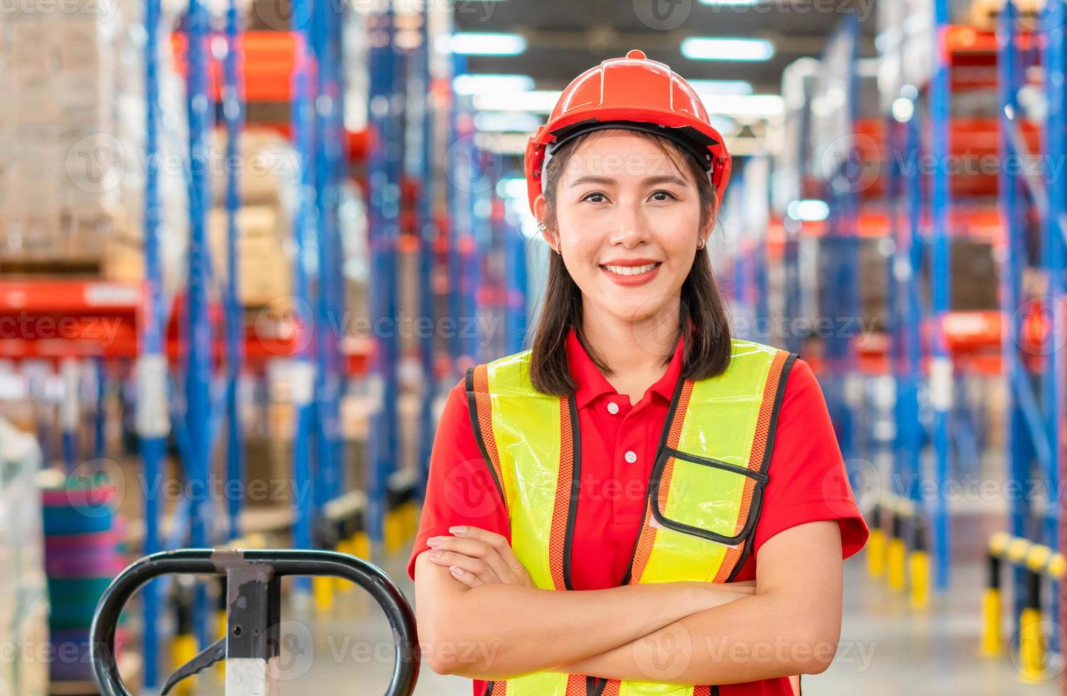 jonge vrouwelijke magazijnmedewerker die in het magazijn staat met gekruiste arm, glimlachende vrouw met een helm die naar camera kijkt met gekruiste armen in het magazijn, werknemer die in het magazijn van de logistieke industrie werkt foto