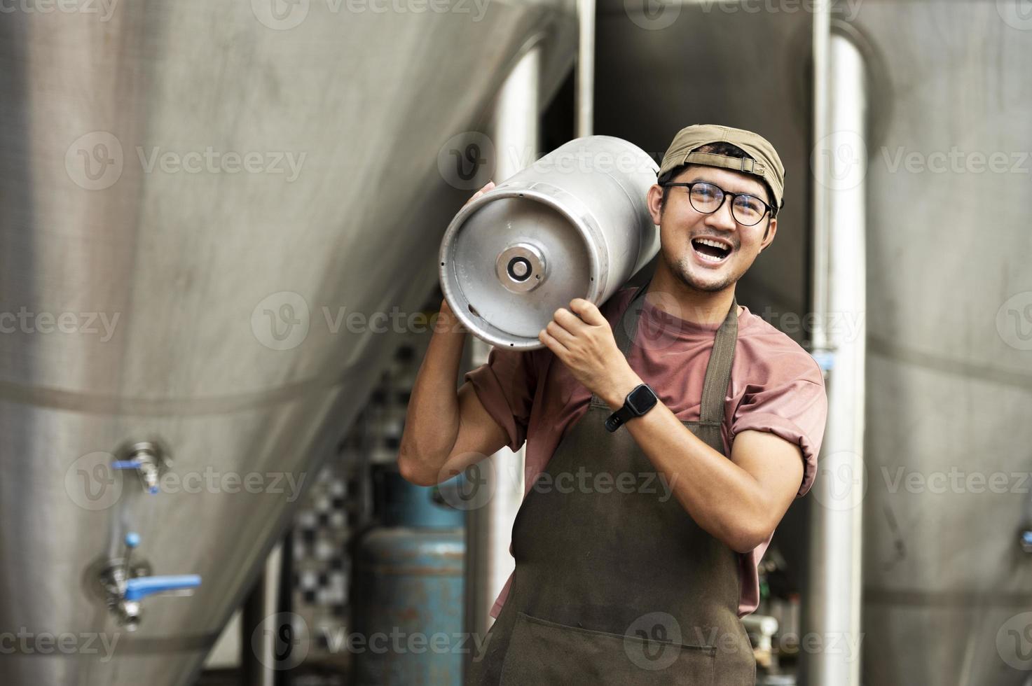 jonge man in leren schort met biervat bij moderne brouwerij, ambachtelijke brouwerijmedewerker foto