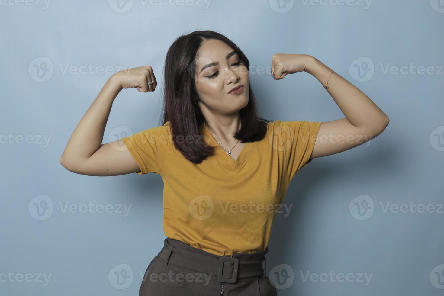 jong mooi meisje draagt een casual geel t-shirt dat over een geïsoleerde blauwe achtergrond staat en de armenspieren trots glimlachen. fitness-concept. foto