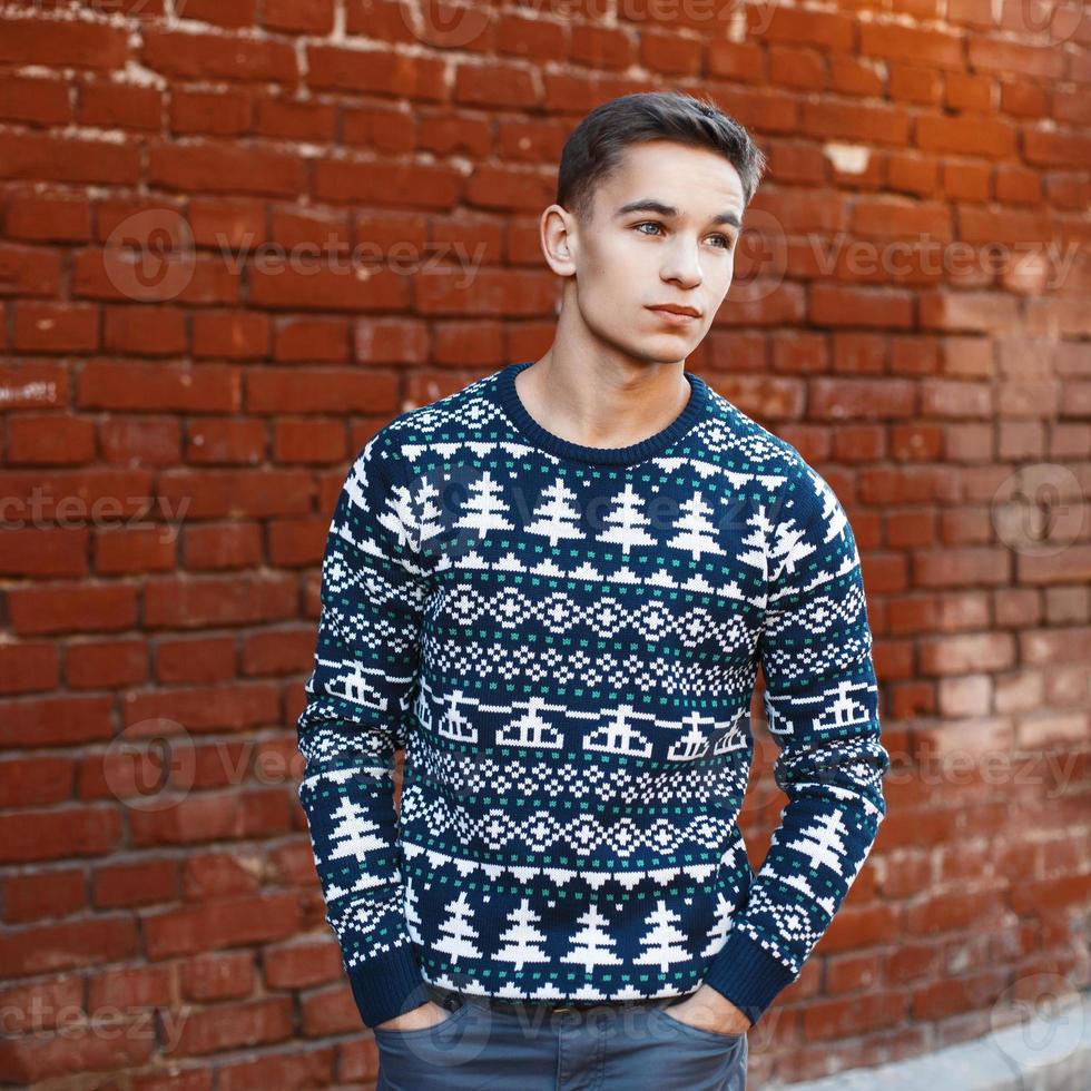 jonge knappe man in een gebreide trui met kerstversieringen op een achtergrond van rode bakstenen muur foto