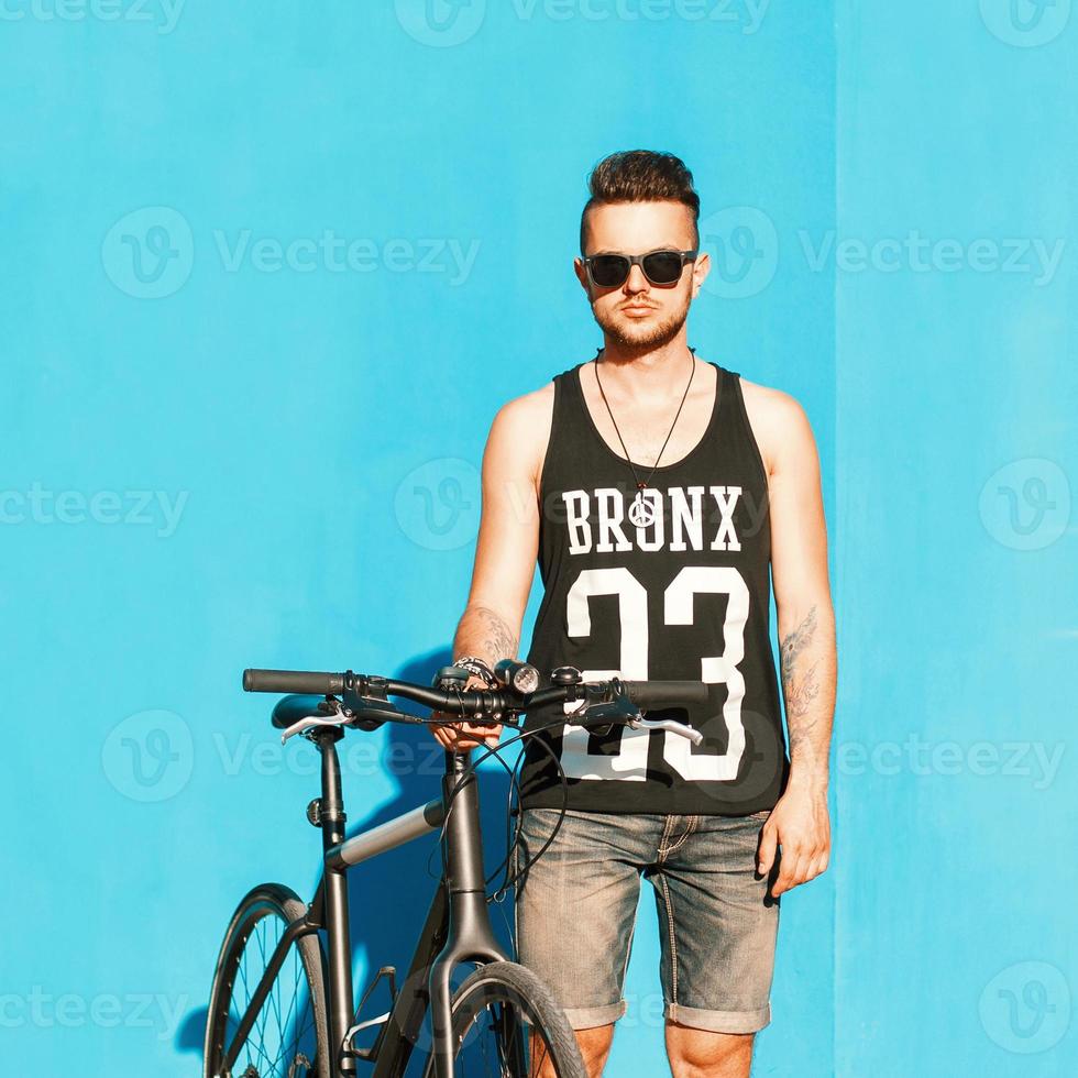 jonge knappe jongen in zonnebril, een zwart t-shirt en denim shorts rijden op een fiets. op de achtergrond van een helderblauwe muur. foto