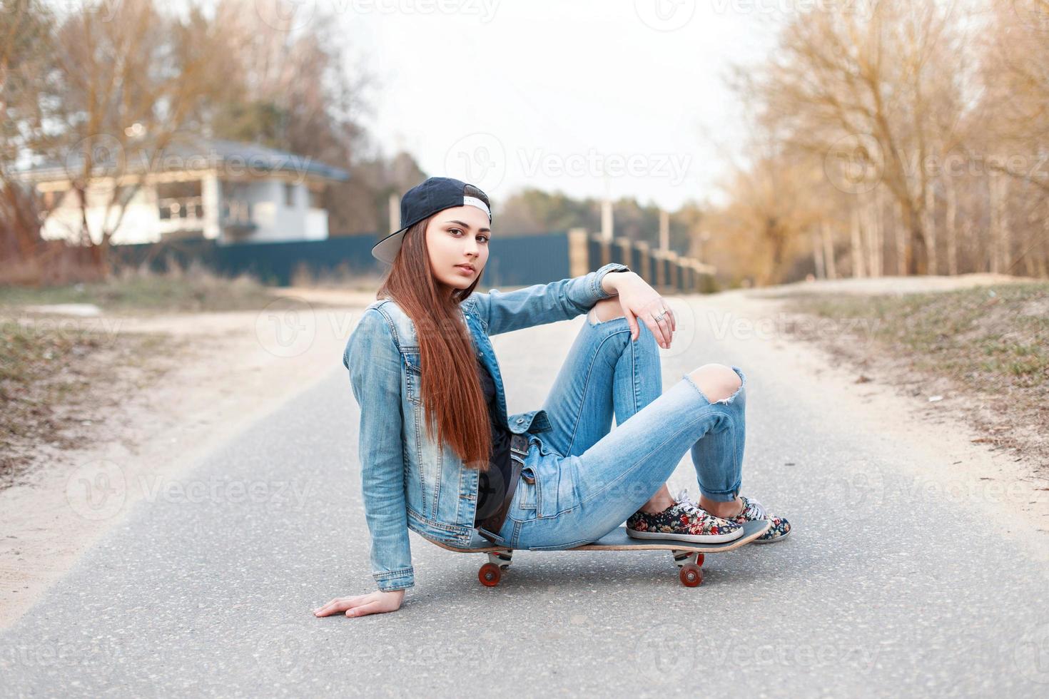 jonge mooie vrouw in een denim jurk en een zwarte baseballcap zit op een skateboard. foto