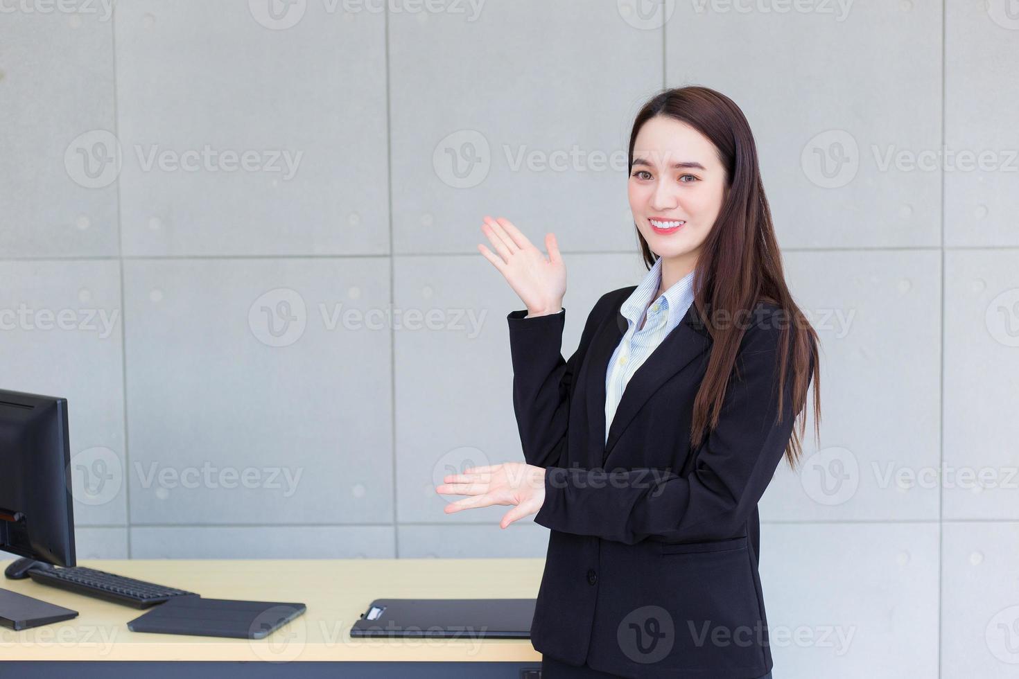 professionele jonge zaken werkende aziatische vrouw die een zwart pak draagt, staat en presenteert iets terwijl ze gelukkig op kantoor als achtergrond glimlacht. foto