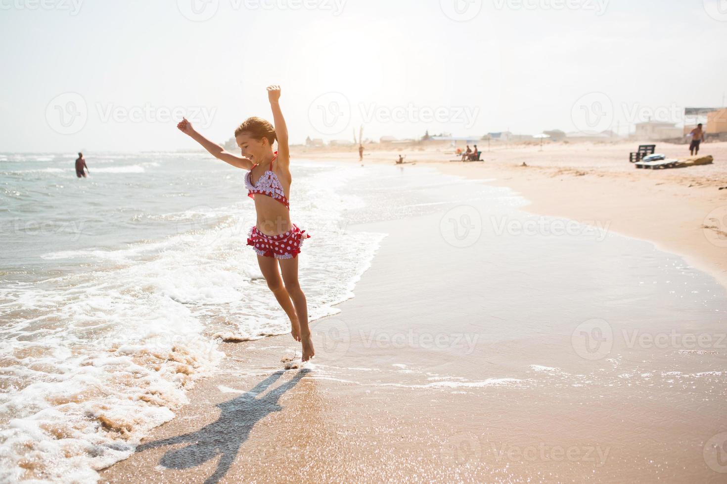 een klein meisje in een rood zwempak speelt op het strand met een zeegolf - springen, rennen, plezier maken. zwemmen, reizen, spelen met water foto