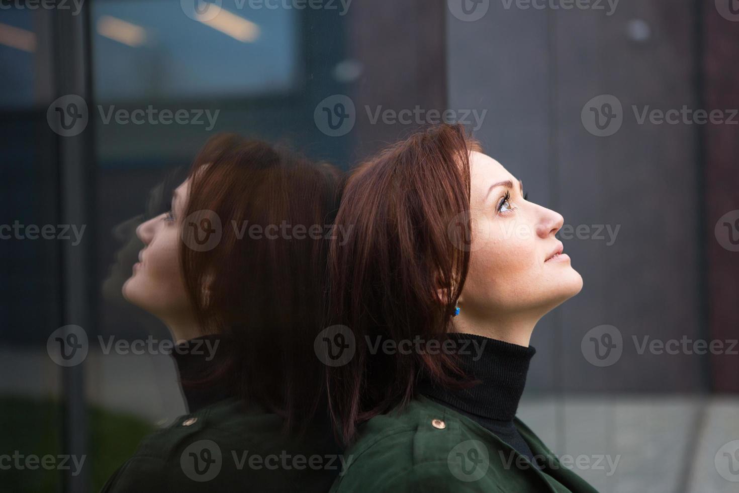 een brunette vrouw met een kort kapsel staat met haar rug naar de spiegelwand, wordt weerspiegeld en kijkt verdrietig. het thema eenzaamheid foto