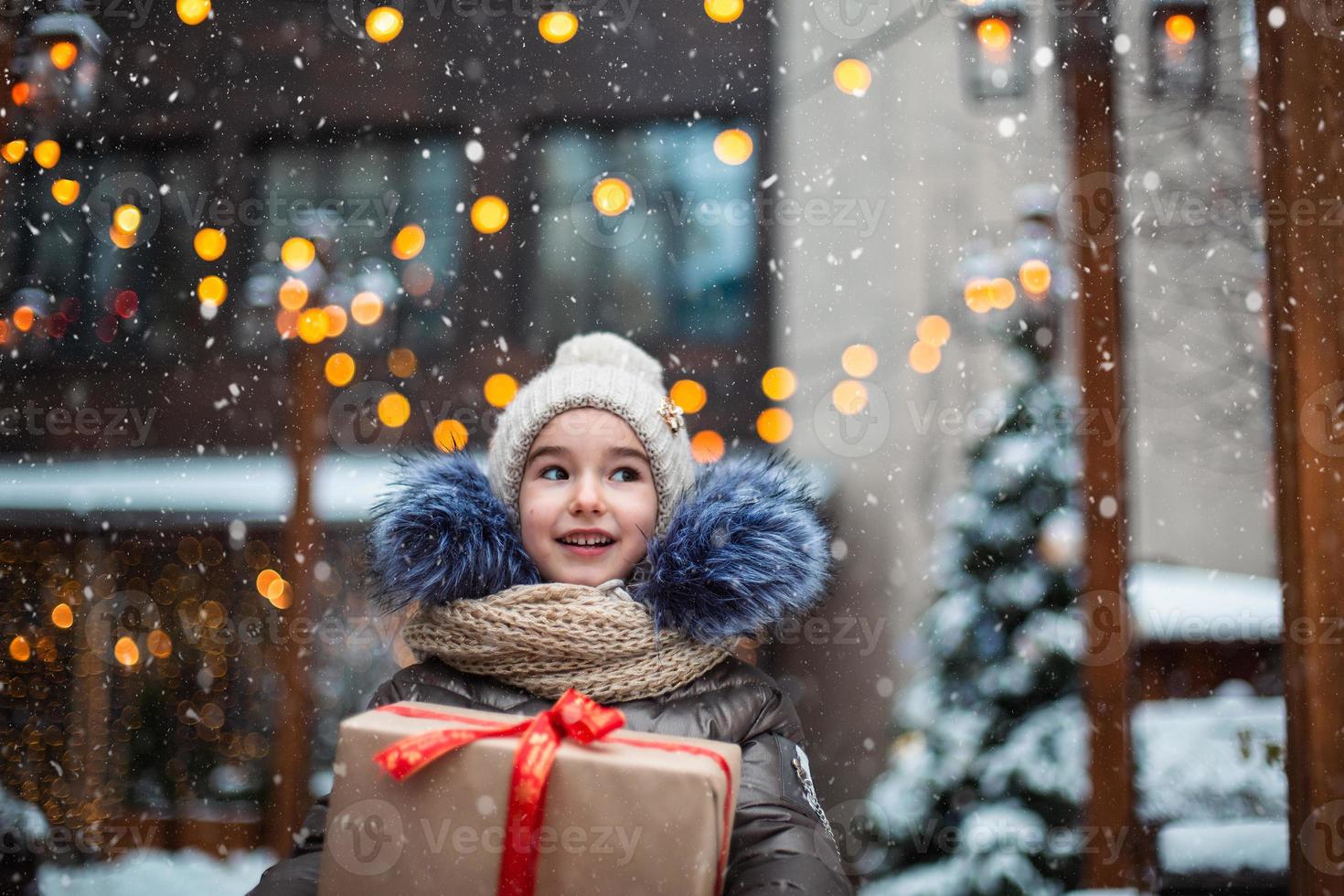 portret van een vrolijk meisje met een geschenkdoos voor kerstmis op een stadsstraat in de winter met sneeuw op een feestelijke markt met versieringen en kerstverlichting. warme kleding, gebreide muts, sjaal en bont. Nieuwjaar foto