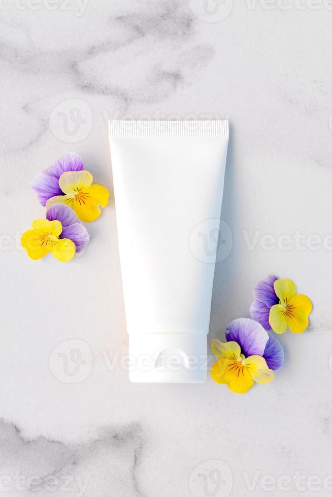 mockup van merkloze witte knijpflesbuis en kleine viooltjesbloemen op marmeren tafel. bovenaanzicht. natuurlijke biologische spa cosmetica concept. leeg voor branding en label. foto