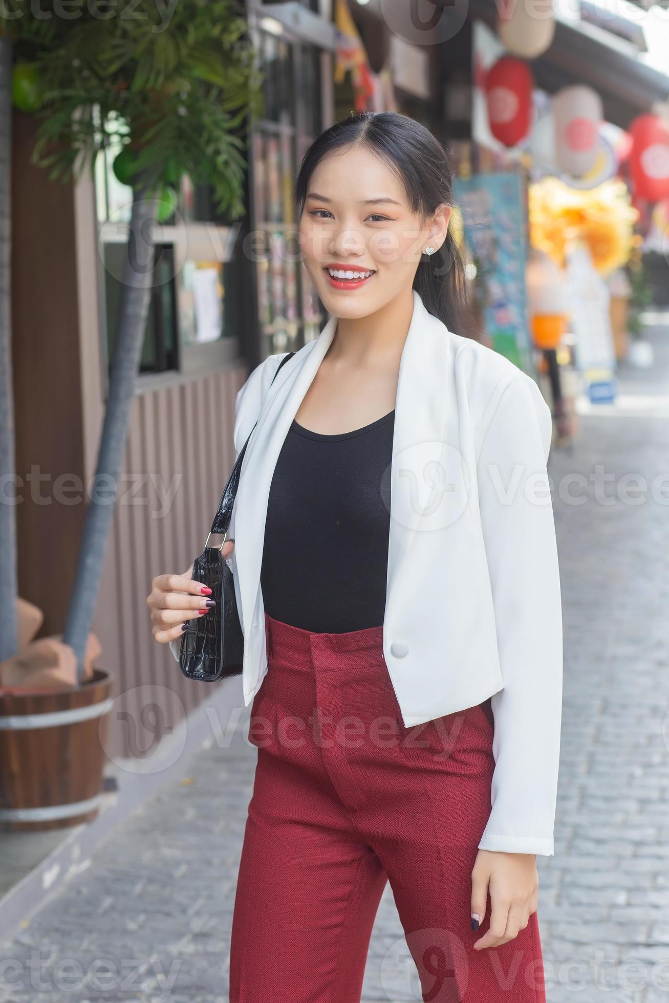 vegetarisch Schurend doorgaan met jong mooi Aziatisch vrouw slijtage wit pak en rood broek lachend, dragen  zak terwijl wandelen in de stad. 17401180 Stockfoto