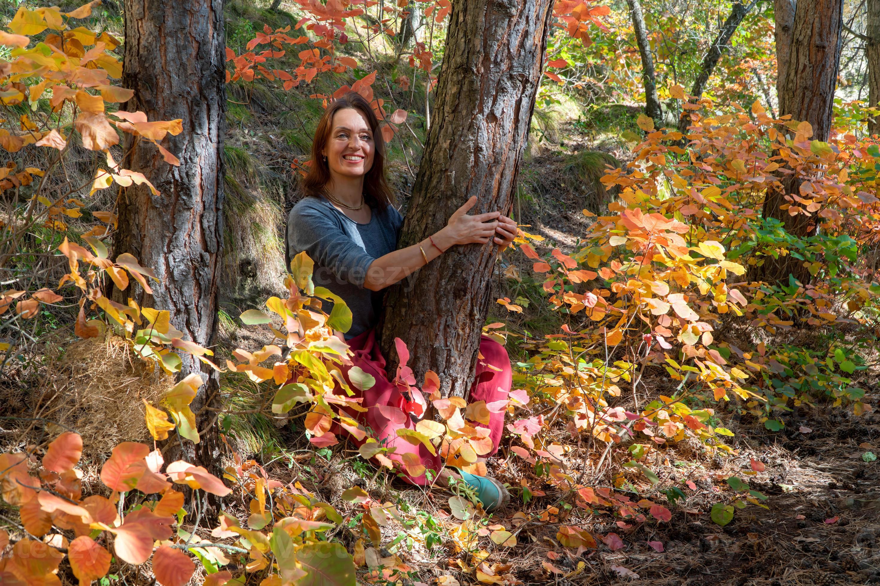 lachende vrouw met rode indische broek zittend in het bos onder de bomen op  het pad met herfstgebladerte. portret van een gelukkige vrouw 5411364  stockfoto bij Vecteezy