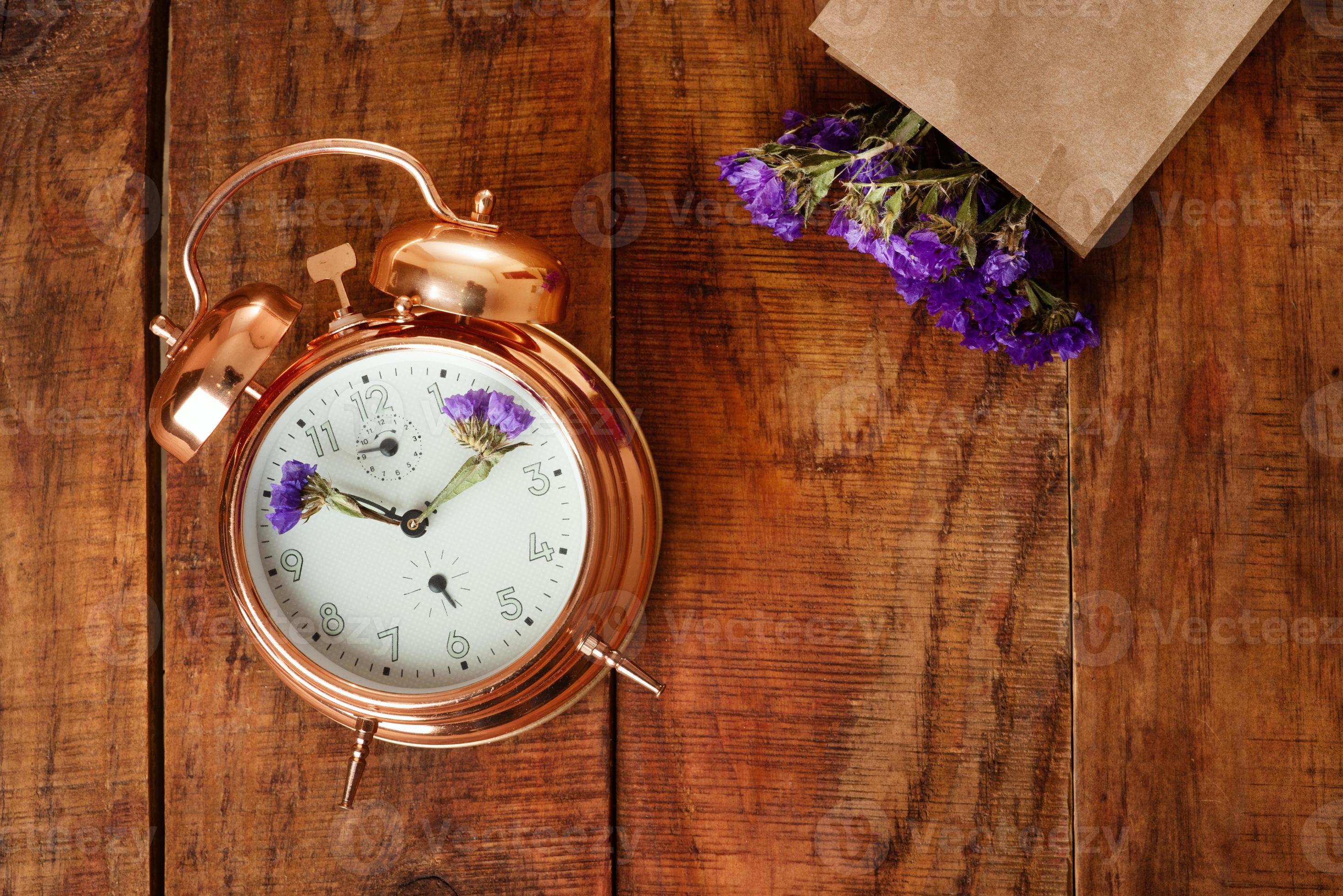 lassen hulp in de huishouding onder gouden wekker met blauwe bloemen in plaats van wijzers en papieren zak met  bloemen op een houten oppervlak. lentetijd 6173764 Stockfoto