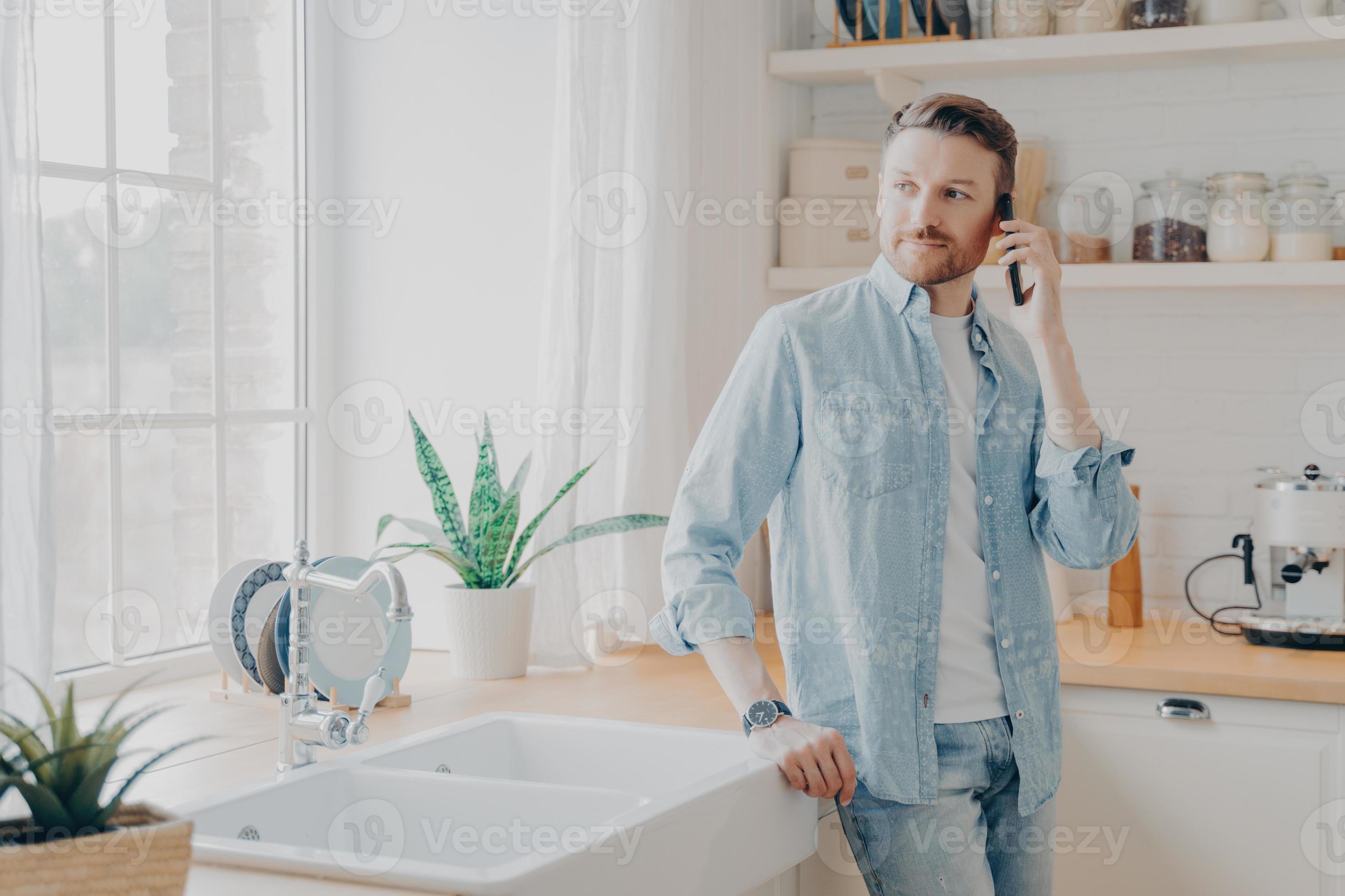 knappe jonge man in casual jeans shirt spreken op mobiel in keuken 8359647