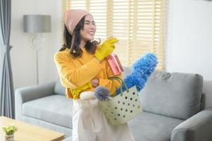 jonge gelukkige vrouw die gele handschoenen draagt en een mand met schoonmaakmiddelen in de woonkamer vasthoudt. foto