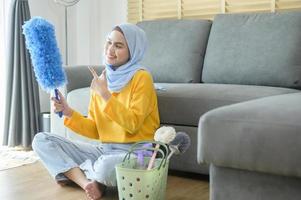 jonge gelukkige moslimvrouw die gele handschoenen draagt en een mand met schoonmaakmiddelen in de woonkamer vasthoudt. foto